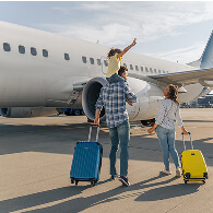 A family with a small child on dad's shoulders rolls their suitcases toward a jet plane for boarding