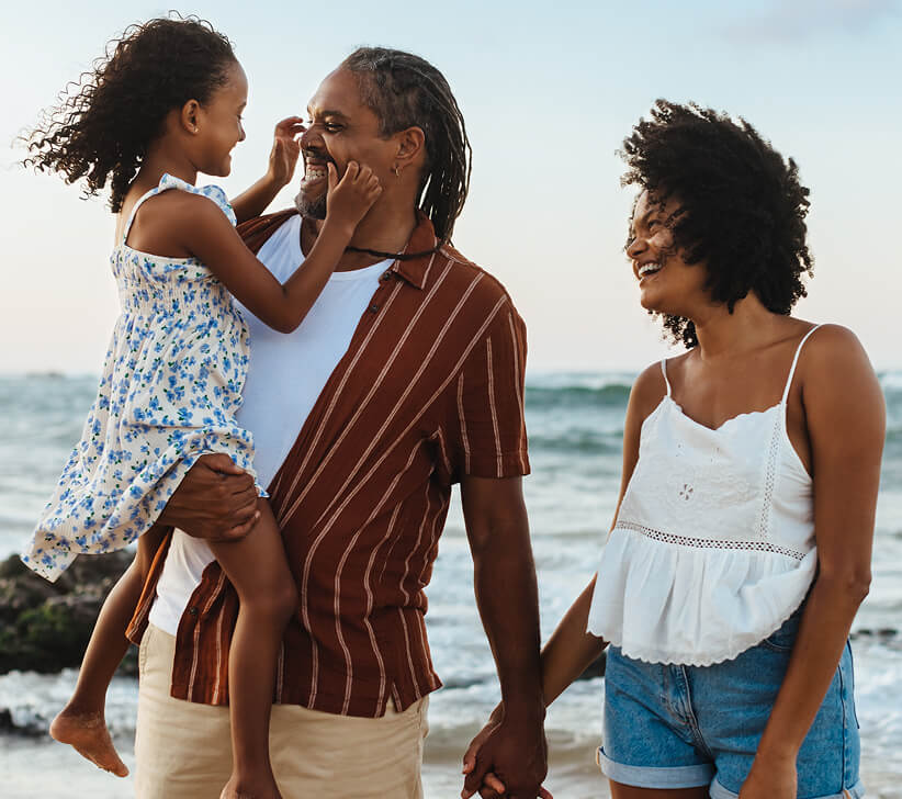 A family at the beach, the father smiling and carrying a young girl while holding his partner's hand.