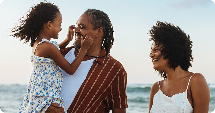 A family at the beach, the father smiling and carrying a young girl while holding his partner's hand.