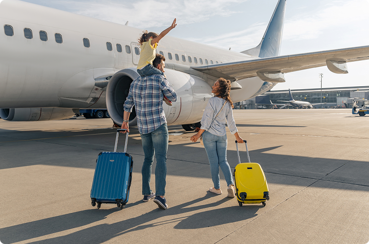 A family with a small child on dad's shoulders rolls their suitcases toward a jet plane for boarding