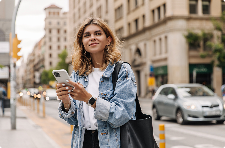 A young woman in a denim jacket looks up from her phone and smiles as she walks in the city center