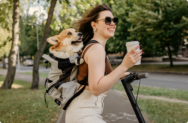 A smiling woman holds a cup of coffee in one hand and rides a bike with a corgi strapped to her back in a pet carrier