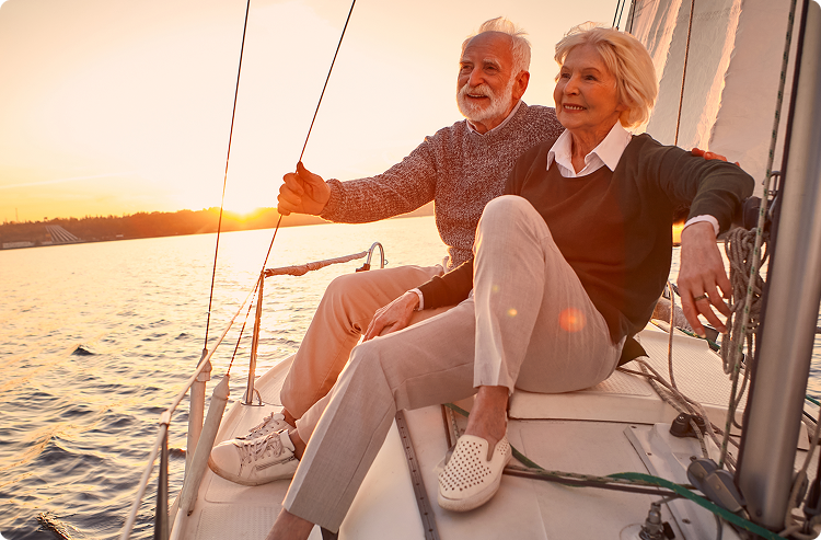 An elderly couple sits on the edge of a sailboat watching the sunset and smiling