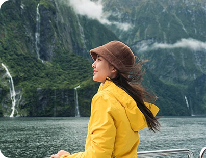 A woman in a yellow rain jacket enjoys scenic mountain and waterfall views from a cruise ship
